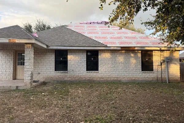 A light brick house with a partially shingled roof and red underlayment showing, with bundles of new shingles on top.