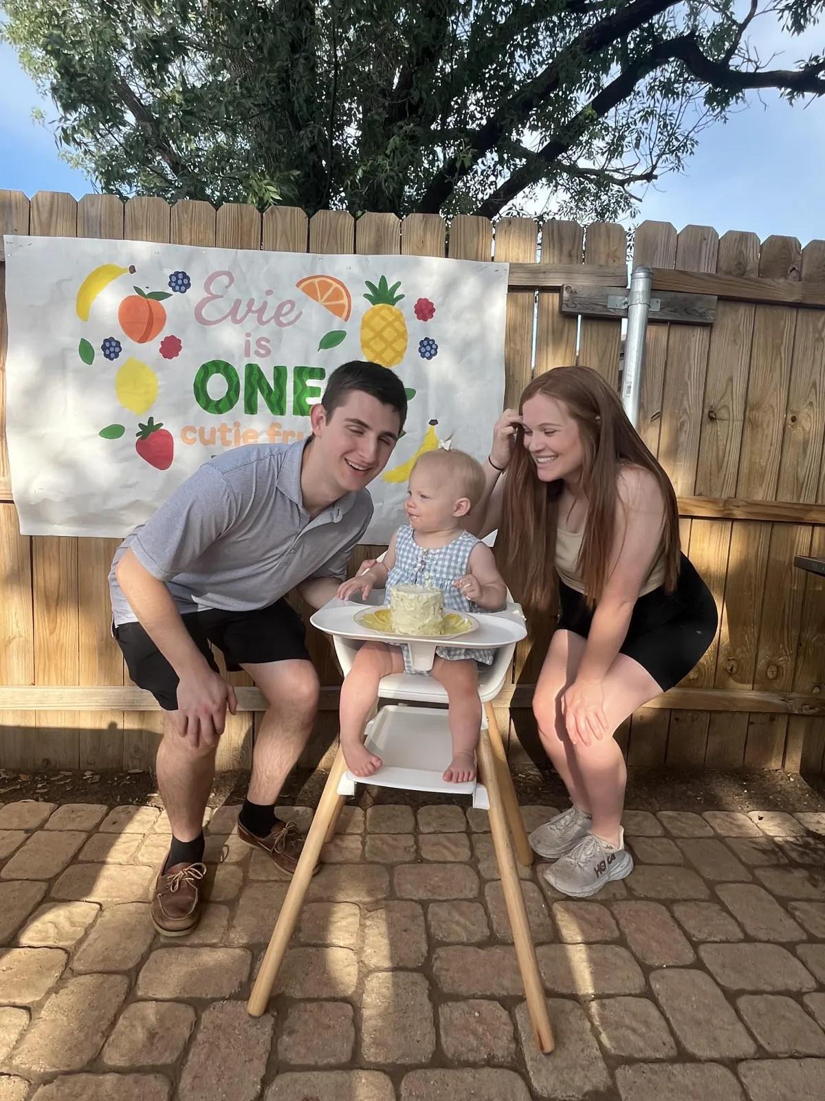 A young family celebrates their daughter's first birthday outdoors. The parents smile and crouch beside their baby, who is seated in a high chair with a small birthday cake. A colorful banner behind them reads "Evie is ONE" with fruit illustrations and the phrase "cutie from the fruitie.