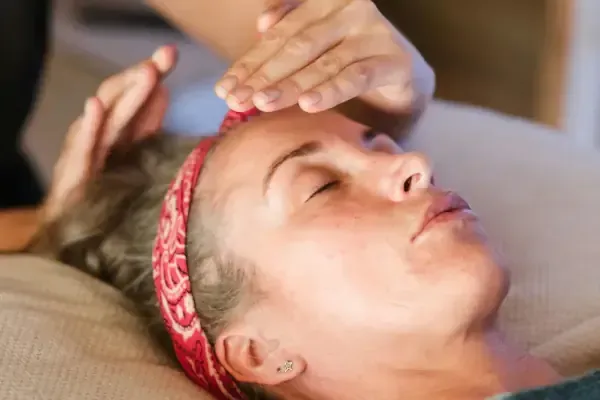 A peaceful client with eyes closed wearing a red patterned headband lies relaxed on a treatment table while a therapist's hands are positioned above their head, performing energy healing or Reiki therapy in a calm, softly-lit treatment room.