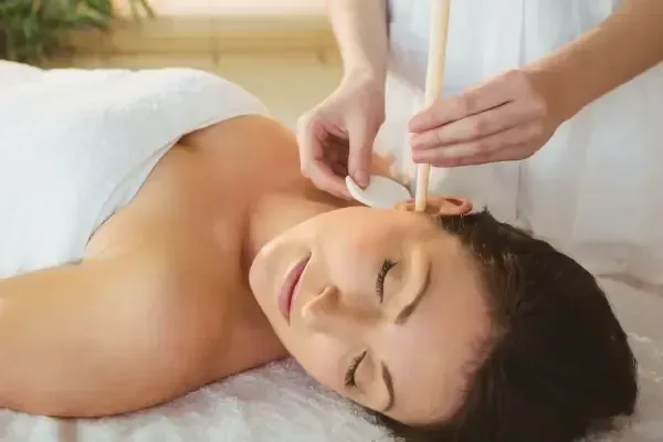 A relaxed client with eyes closed receiving ear candling treatment while lying on a white towel. A therapist's hands guide a lit ear candle near the client's ear in a peaceful spa setting with soft natural lighting.