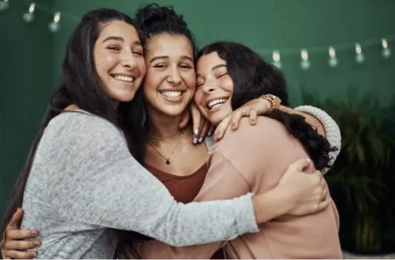 A candid moment of a woman in her 30s, eyes closed and smiling, hands over heart, surrounded by soft peach and rose tones. The background is blurred, suggesting a supportive group setting, with gentle sunlight streaming in, conveying warmth and emotional resonance.