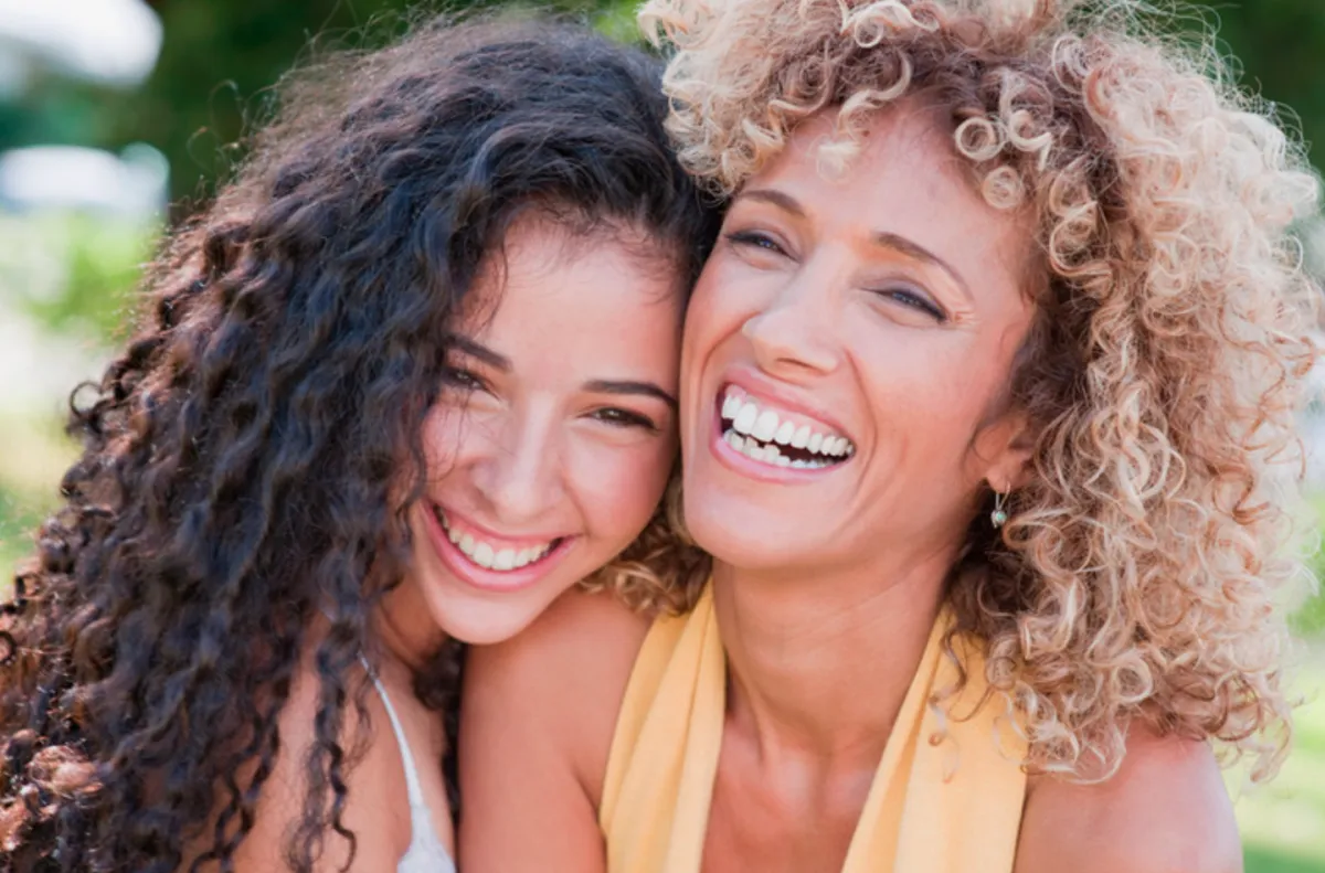A candid moment of a woman in her 30s, eyes closed and smiling, hands over heart, surrounded by soft peach and rose tones. The background is blurred, suggesting a supportive group setting, with gentle sunlight streaming in, conveying warmth and emotional resonance.