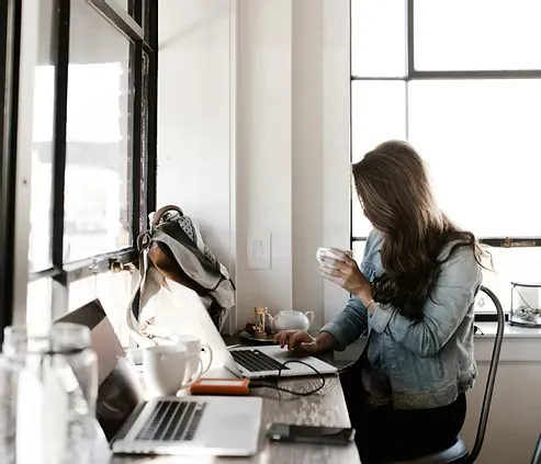 Woman working on a laptop at a bright café or home workspace while holding a cup.