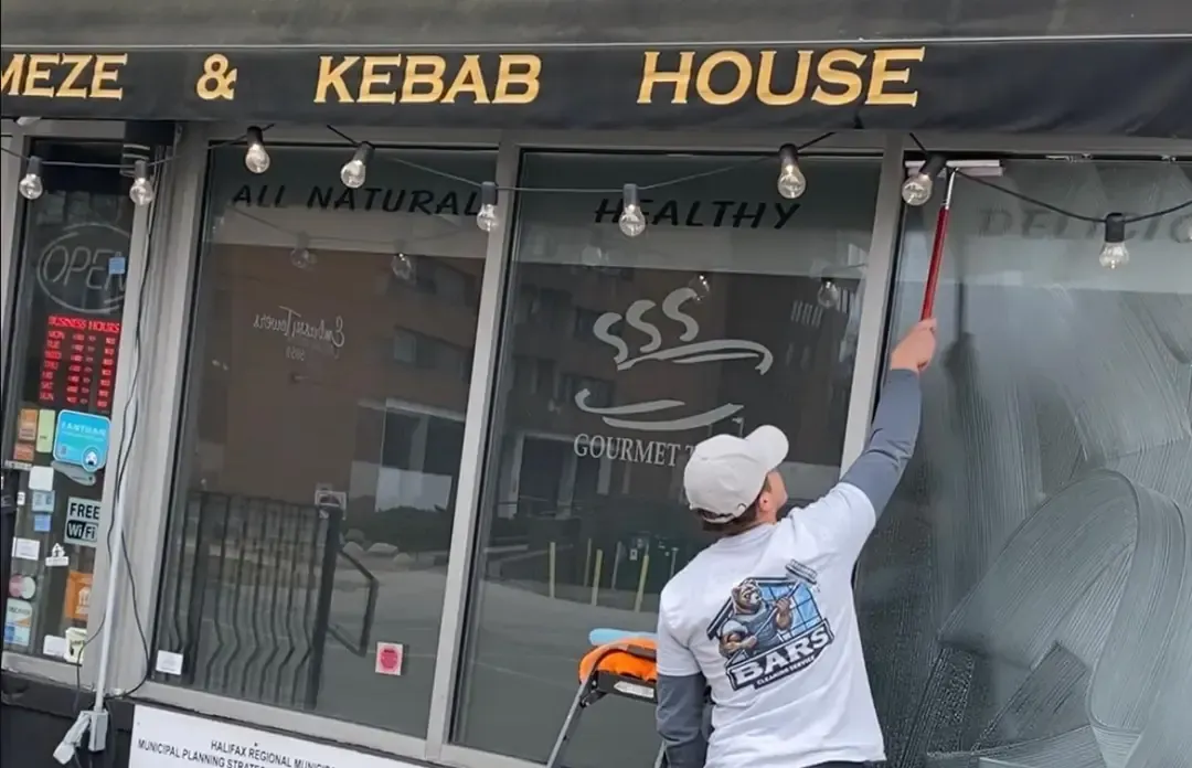 Worker cleaning restaurant windows under a black awning that reads ‘Kebab House,’ wearing a Bars Property Services shirt.