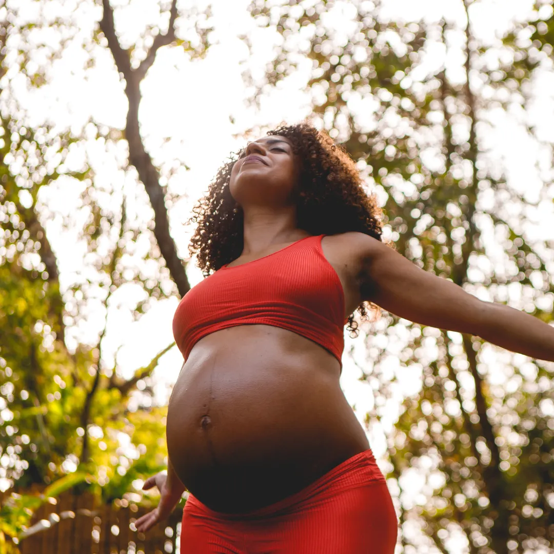 A pregnant woman dressed in red stands in the woods, embodying a peaceful moment amidst nature's beauty.