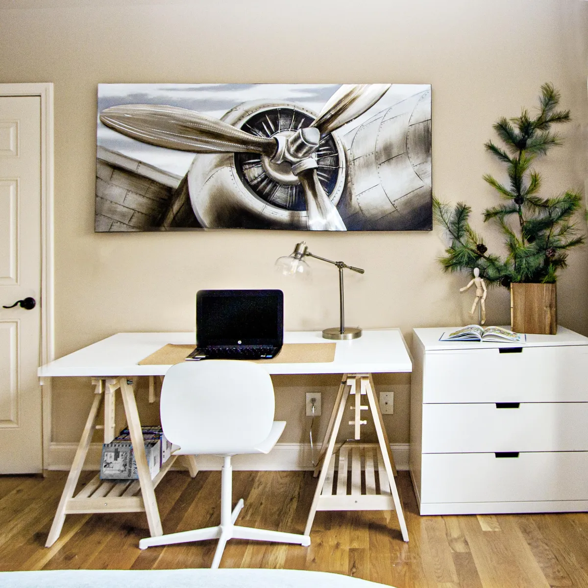 White wooden desk with matching chair, laptop, mini tree, and wall art of a plane.