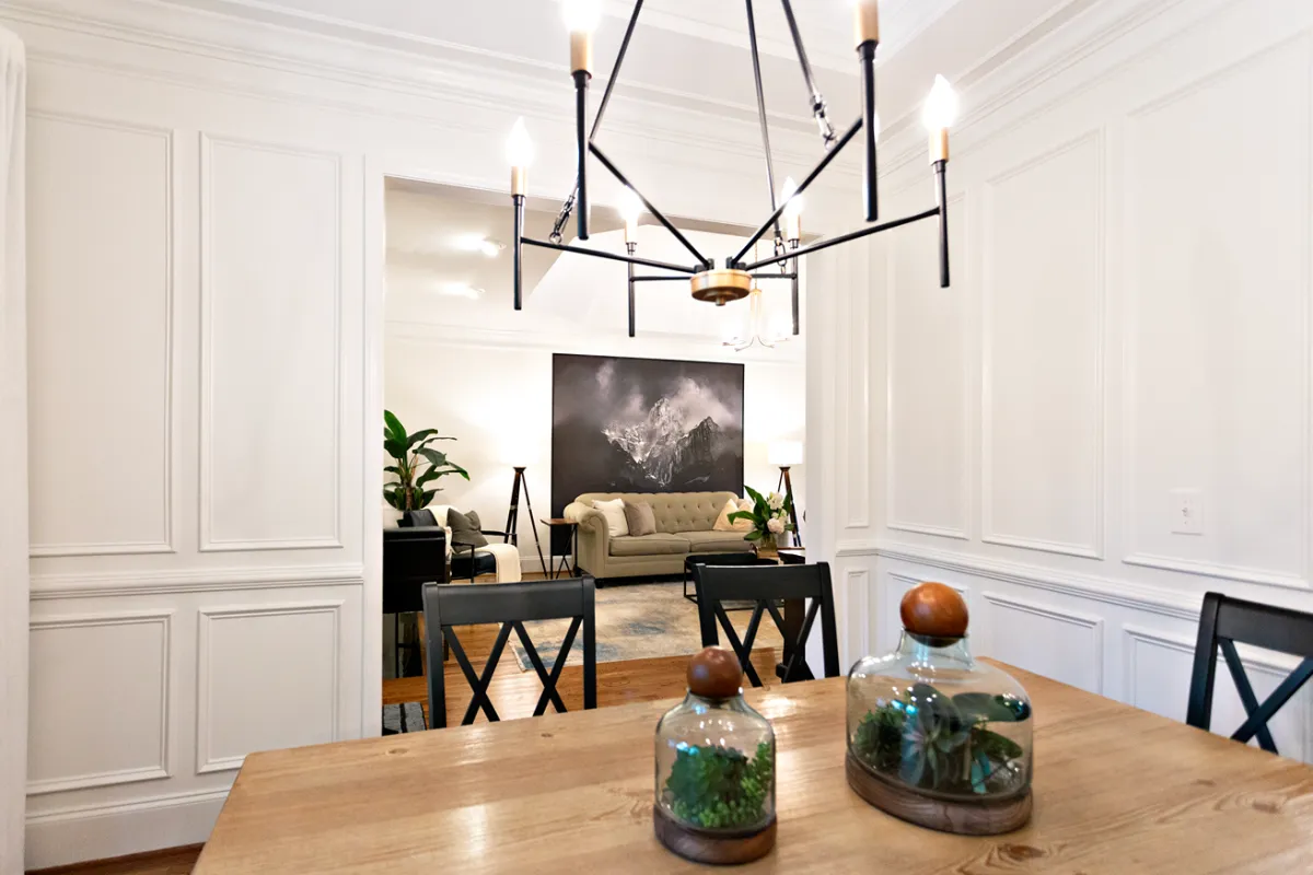 Traditional styled dining room with a connected living room in the background, showcasing white walls with traditional panel moldings, and a close-up of the dark metal chandelier.