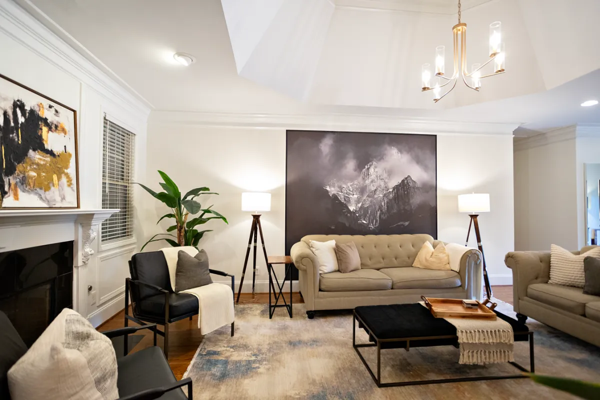 Traditional earthy-toned living room with fireplace, matching black armchairs, linen couches, and a cushioned foot rest.