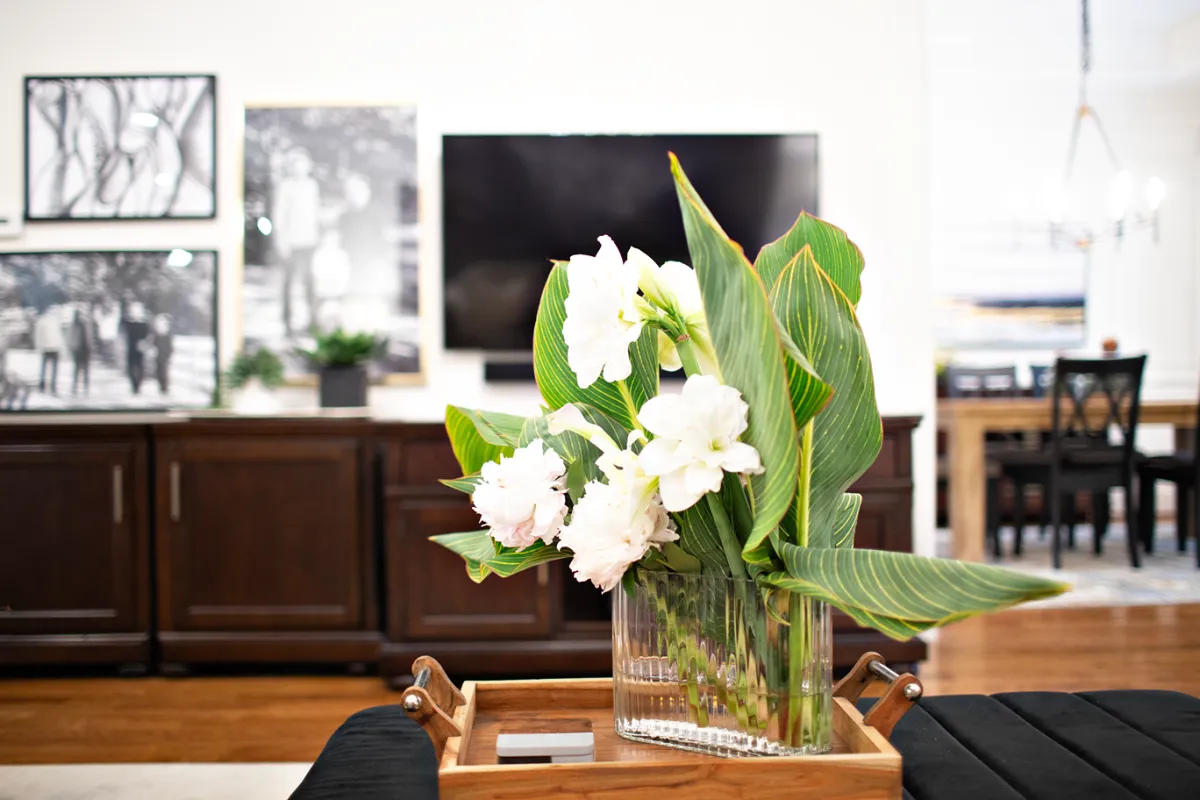 Cushioned footrest with a wooden tray strategically placed on it, with a glass vase of white flowers and large green leaves.