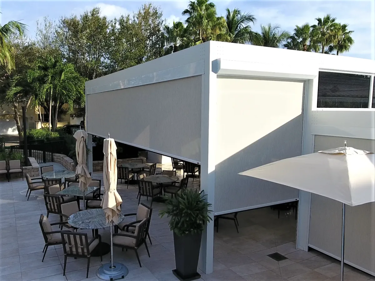 An outdoor patio area with tables and chairs under opened umbrellas beside a modern white architectural structure with large windows and landscaped bushes, equipped with retractable bug screens to protect your guests.