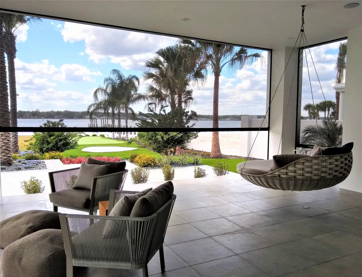 Modern living room with large glass windows featuring retractable insect screens, overlooking a lake, and equipped with a hanging chair, sofa, and striped chair, with palm trees visible outside.