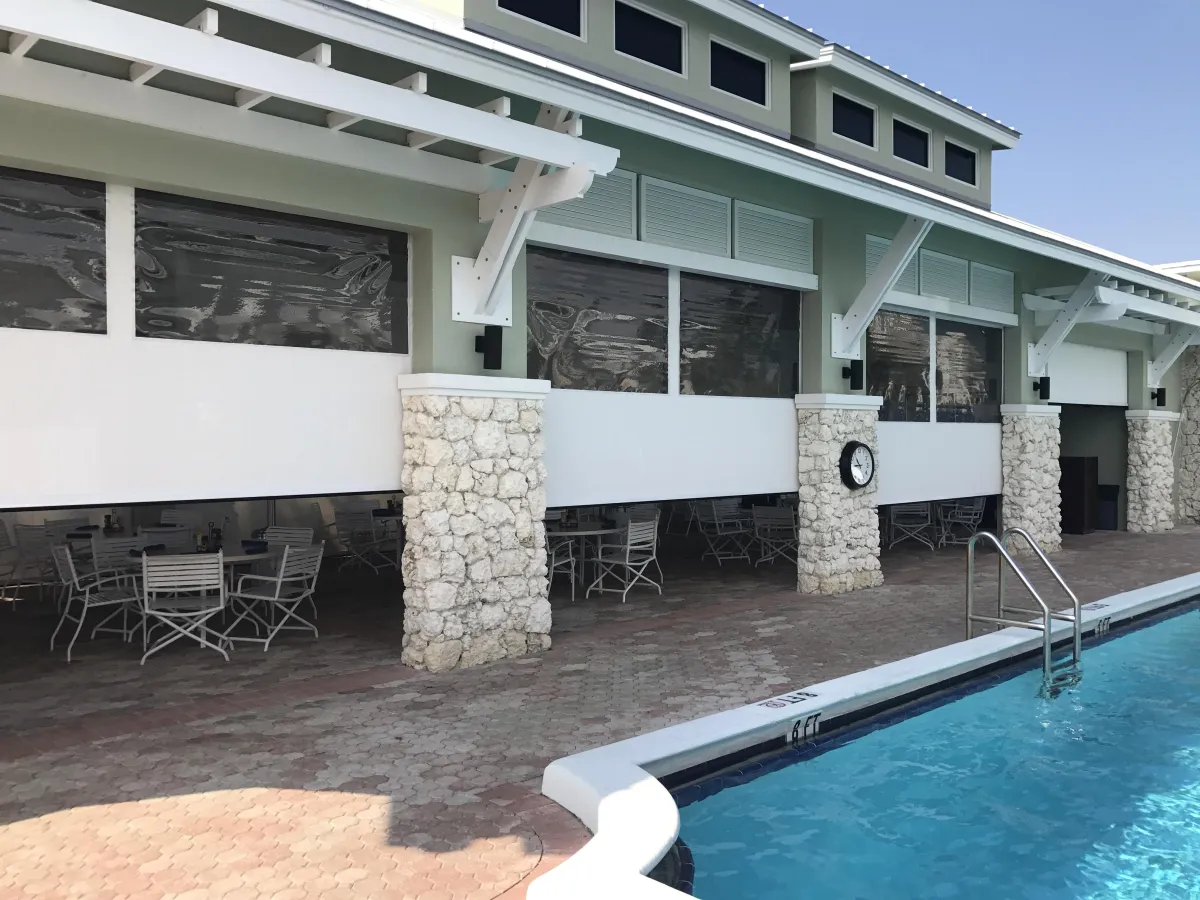 A modern beach house with white walls and stone columns, large windows equipped with motorized retractable screens, adjacent to a swimming pool with lounge chairs and a paved deck.