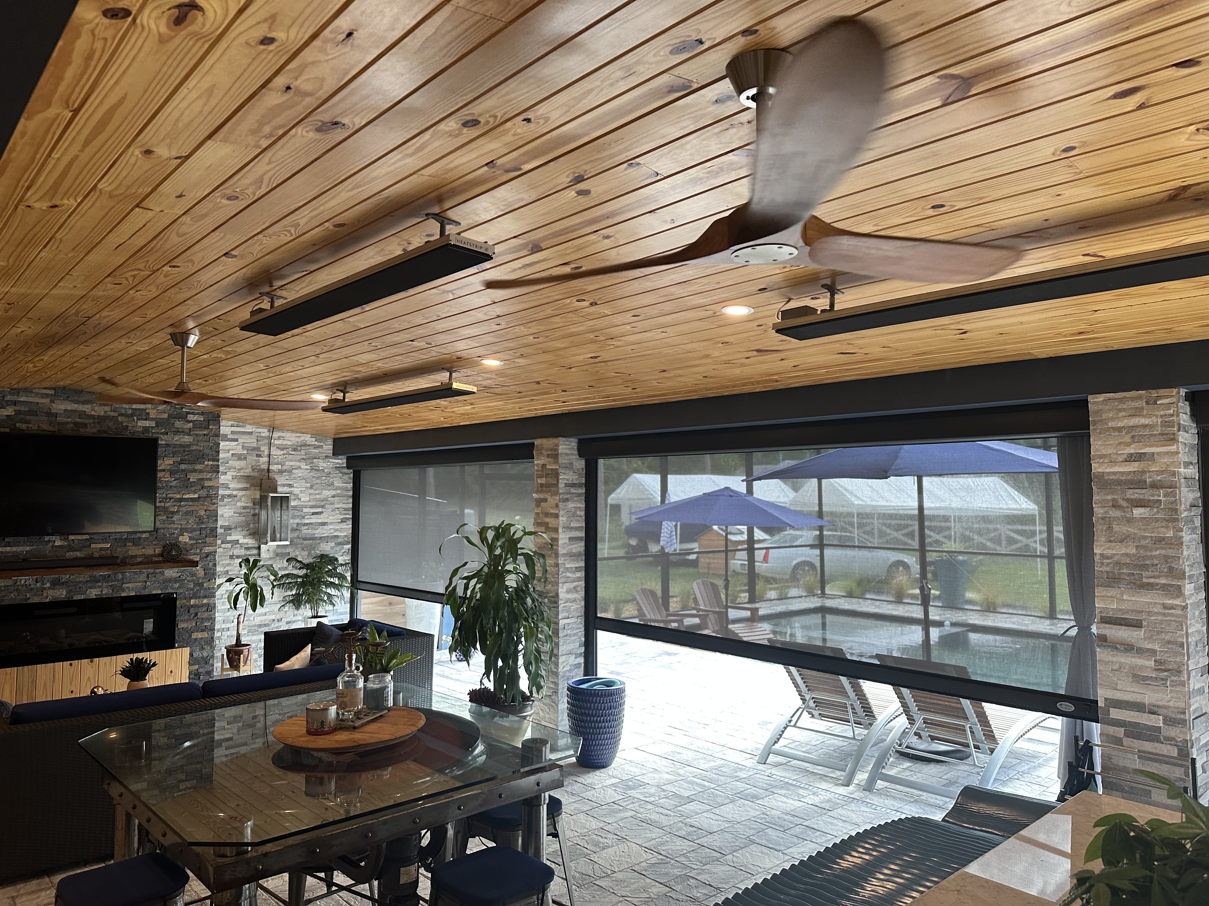 A well-kept backyard with a spacious covered patio featuring retractable Defender Motorized screens attached to a modern house, showcasing white pillars and a lush green lawn under a clear blue sky.