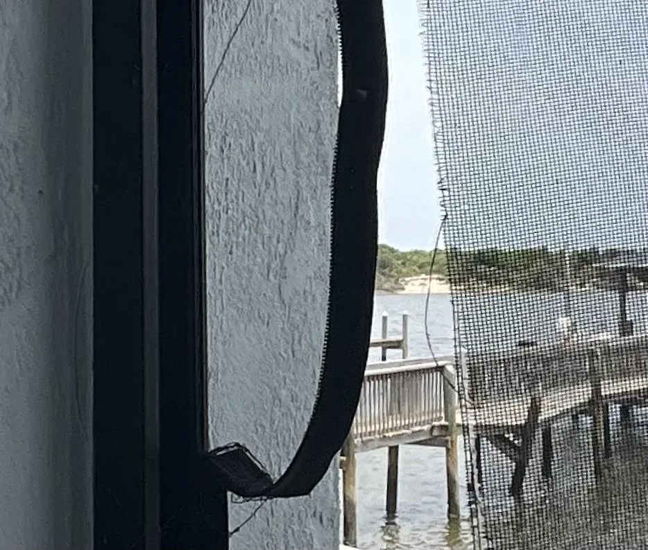 A view through a window corner shows a ripped zipper track screen with a partial view of a dock and water body in the background, framed by a light-colored wall and black piping.