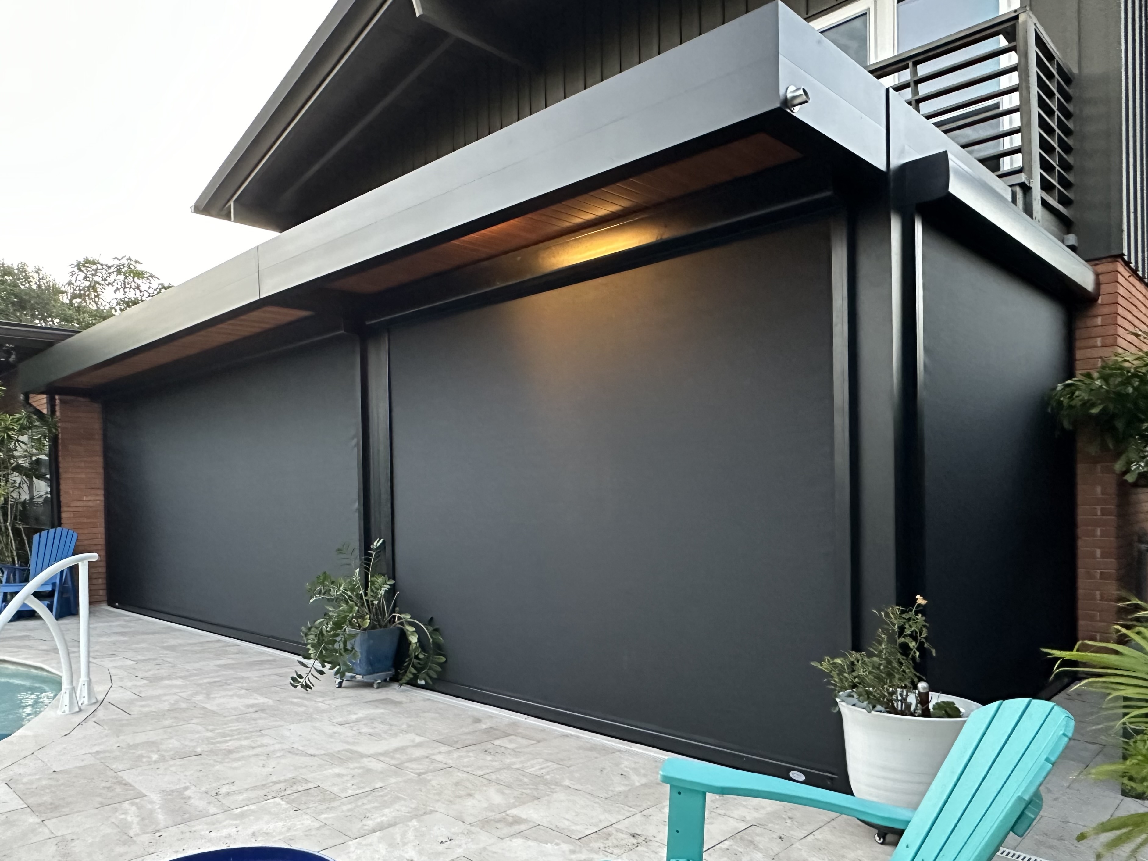 Outdoor seating is beneath a pergola area with a large, modern, hassle-free, black retractable awning extending over a patio beside a swimming pool. Blue and green chairs add a splash of color. Picture of a white house with black MaganaTrack motorized screen deployed. 