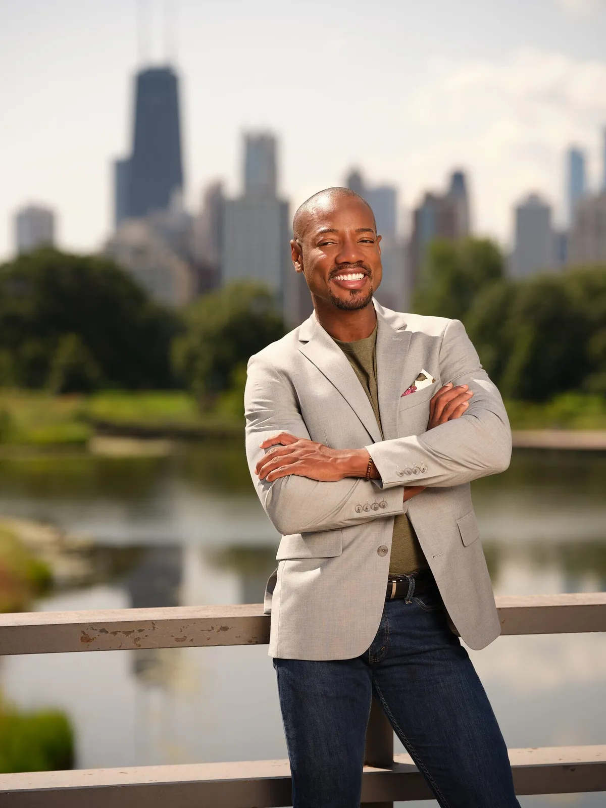 Smiling man in a beige blazer with arms crossed, standing in front of a lake and a city skyline