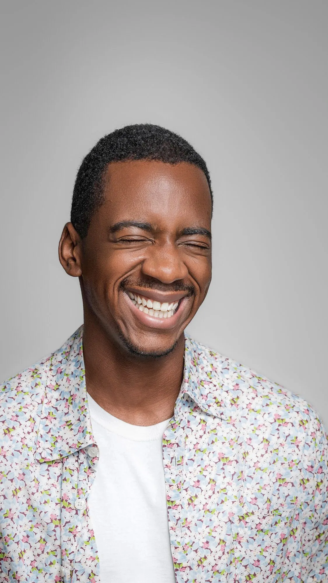 Headshot on pale grey, a floral shirt and a wide laugh