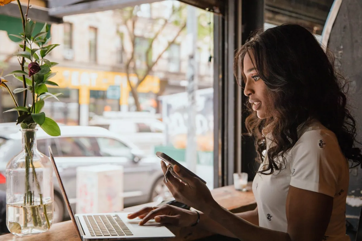 Young professional woman working remotely in a cafe, using laptop and smartphone by the window with city view.