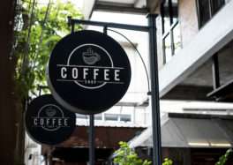 Modern black circular hanging signboard for a coffee shop with steaming cup logo and bold 'COFFEE SHOP' text, displayed outdoors during daytime.