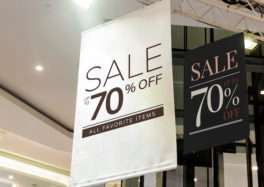 Indoor view of two promotional hanging signs promoting a major sale: a tall white banner stating 'SALE UP TO 70% OFF ALL FAVORITE ITEMS' and a black banner stating 'SALE UP TO 70% OFF', suspended from the ceiling near glass doors with bright overhead lighting.