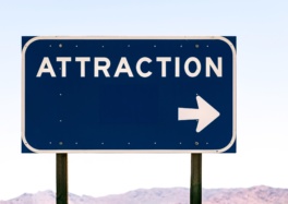 Tourist or directional road sign in dark blue with bold white lettering spelling 'ATTRACTION' and a white arrow indicating direction to the right, mounted against a pale sky background