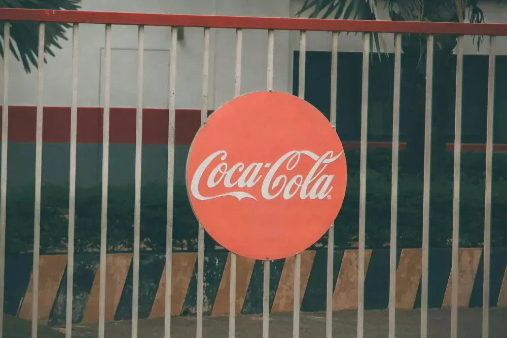 Branded circular Coca Cola sign mounted on an exterior fence, used as an example of commercial signage, brand marketing displays, and outdoor advertising signage.