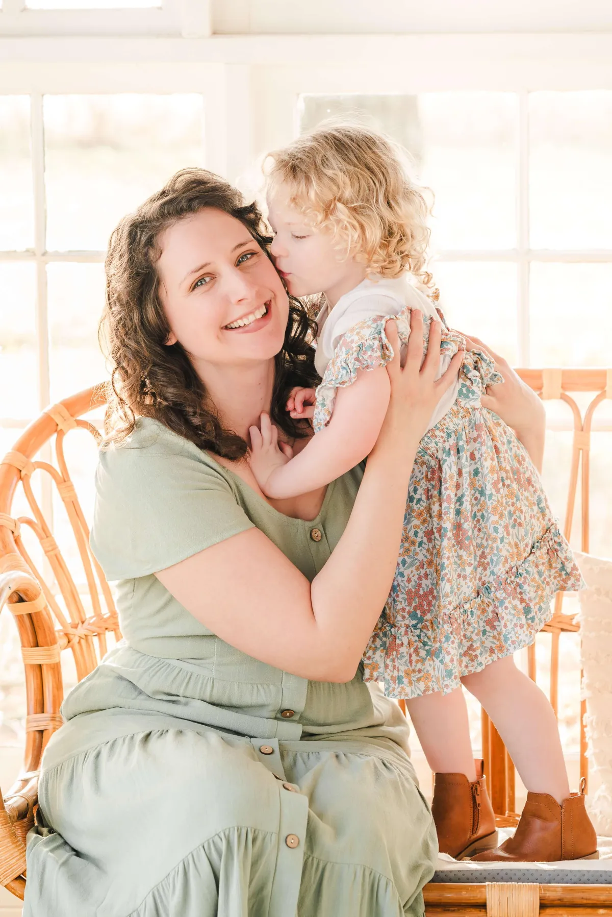 Justine, the photographer at Justine Renee Photography, sits on a bamboo bench. Her young daughter stands on the bench next to her and kisses her on the cheek.