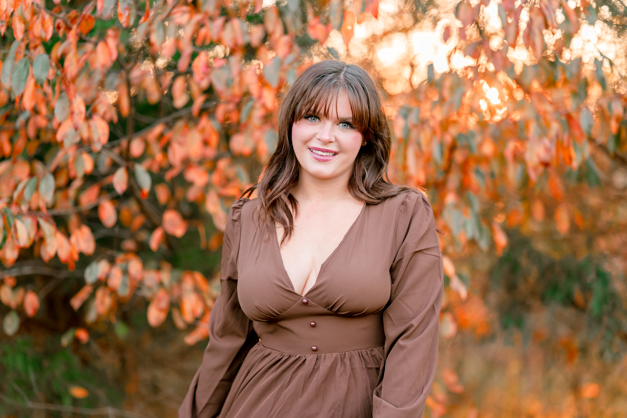 A high school senior in a brown dress poses in front of a tree with orange leaves and the sun peeking through. It's an example of how pretty Bells Mill Park is in the fall.