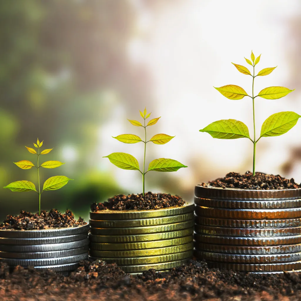 Two smiling women wearing matching purple dresses stand closely together in front of a lush green backdrop of hanging foliage. They appear to be middle-aged, with one having gray hair and the other dark brown hair. Both have bright expressions and are looking at the camera. The natural lighting highlights their faces and the texture of the leaves behind them.
