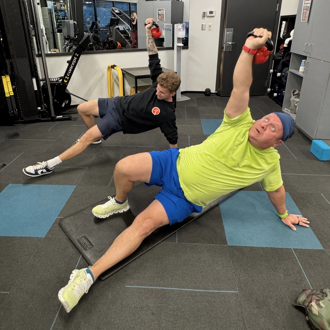 Adults over 40 performing a kettlebell-supported mobility and strength exercise with a personal trainer at Quinntessential Fitness, focusing on core stability, coordination, and controlled movement.