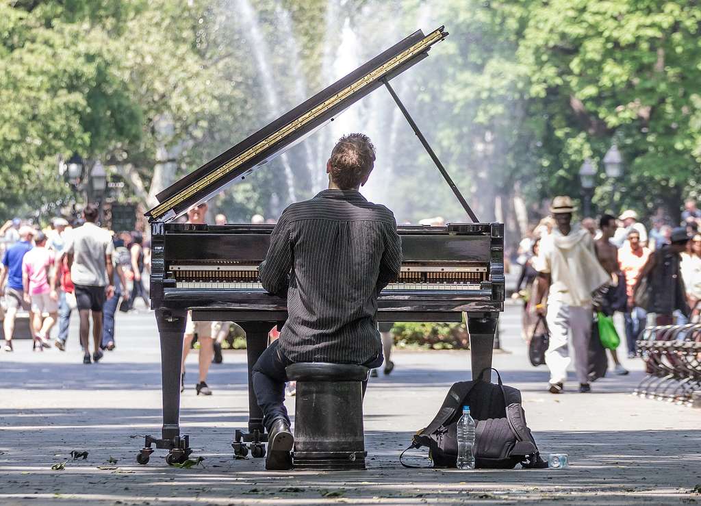 Street pianist playing a grand piano outdoors in a city park while pedestrians walk nearby