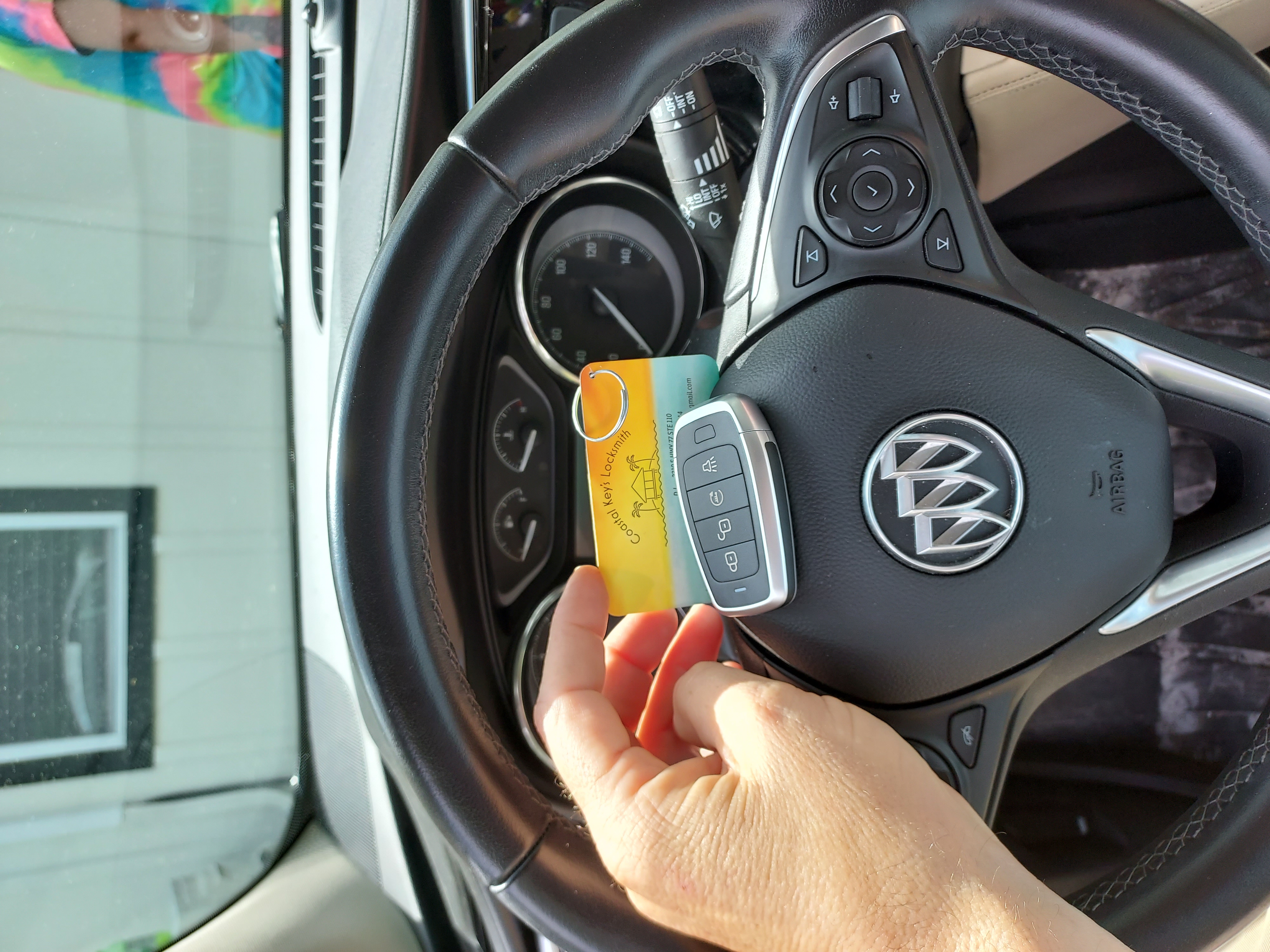 A close-up of a locksmith's hands programming a new car key fob using a digital device inside a modern vehicle. The dashboard and steering wheel are visible, highlighting advanced technology and precision work.