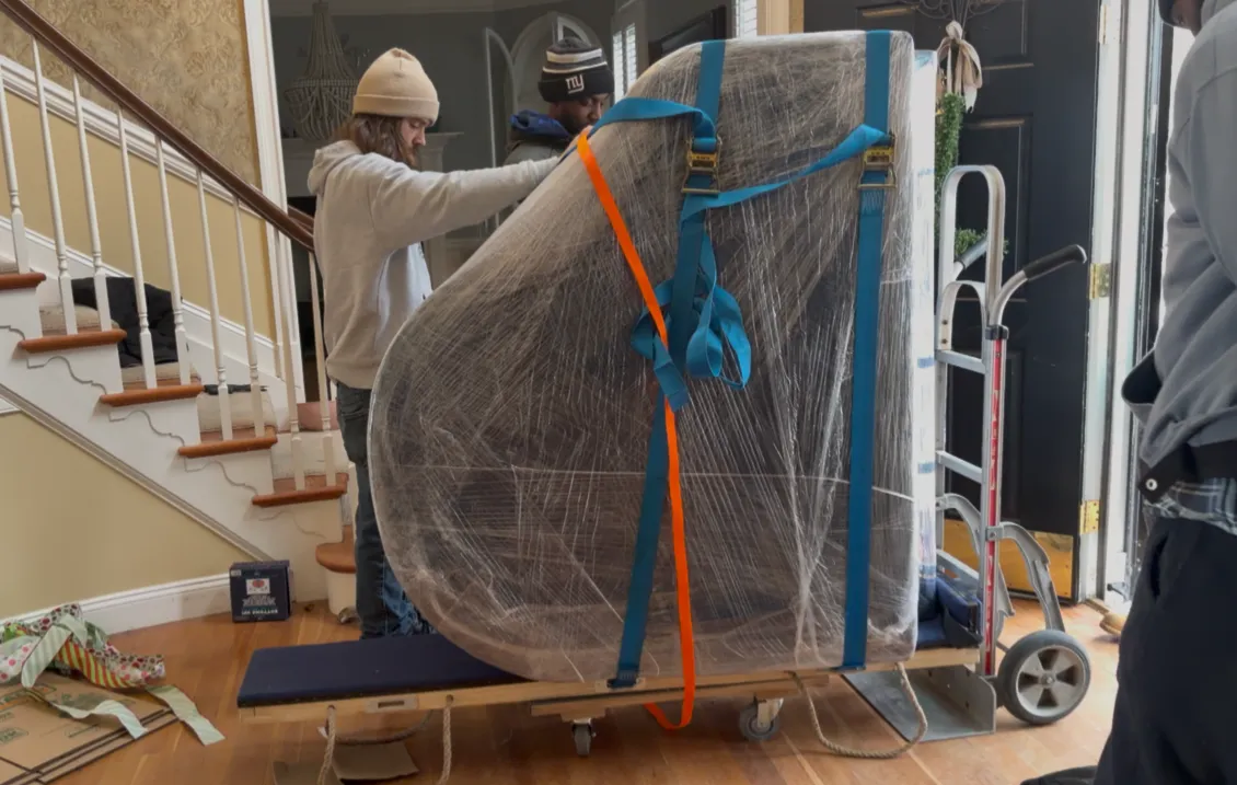 Professional movers safely transporting a large specialty item using protective padding, straps, and moving equipment inside a residence.
