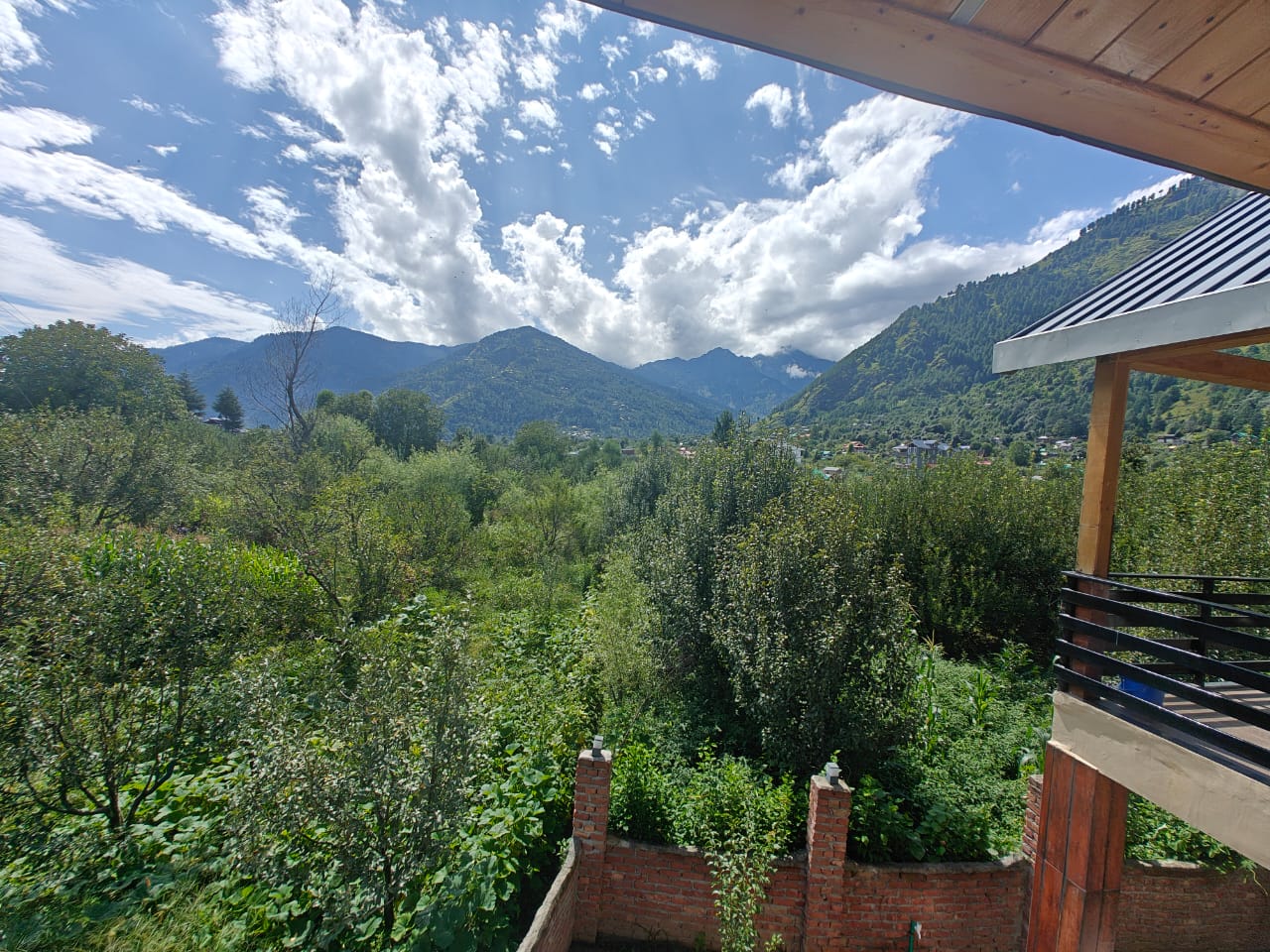 A panoramic exterior shot of a luxury cottage in Manali, surrounded by lush greenery and snow-capped mountains in the distance.