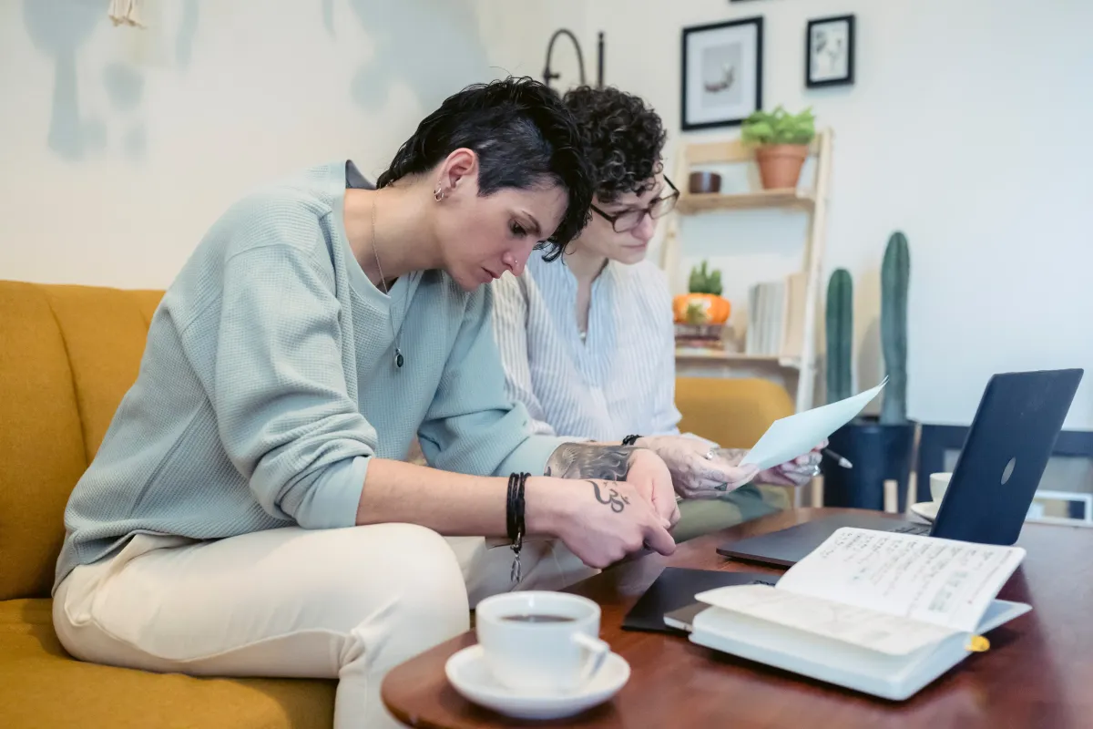 Two women at a desk with a computer and notepad open on it.