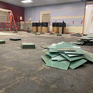 A basement with typical contractor materials strung about and several dozen large floor tiles removed and laying in piles with trash bins in the bakground