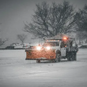 A snow plow truck on an empty road with a red plow on the front of the truck and it's headlights on as the weather appears to be snowing.