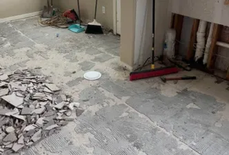 A basement with typical contractor materials strung about and several dozen large floor tiles removed and laying in piles with trash bins in the bakground