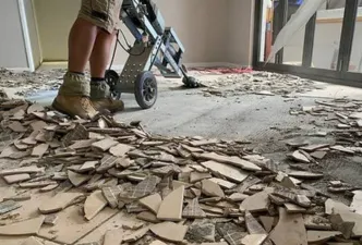 A basement with typical contractor materials strung about and several dozen large floor tiles removed and laying in piles with trash bins in the bakground