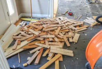 A basement with typical contractor materials strung about and several dozen large floor tiles removed and laying in piles with trash bins in the bakground