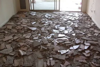 A basement with typical contractor materials strung about and several dozen large floor tiles removed and laying in piles with trash bins in the bakground