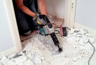 A basement with typical contractor materials strung about and several dozen large floor tiles removed and laying in piles with trash bins in the bakground