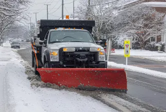 A snow plow truck on an empty road with a red plow on the front of the truck and it's headlights on as the weather appears to be snowing.