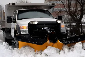 A snow plow truck on an empty road with a red plow on the front of the truck and it's headlights on as the weather appears to be snowing.