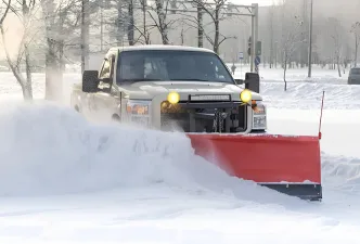 A snow plow truck on an empty road with a red plow on the front of the truck and it's headlights on as the weather appears to be snowing.