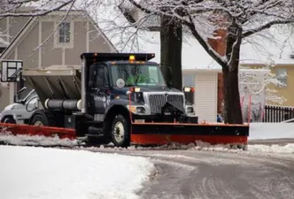 A snow plow truck on an empty road with a red plow on the front of the truck and it's headlights on as the weather appears to be snowing.