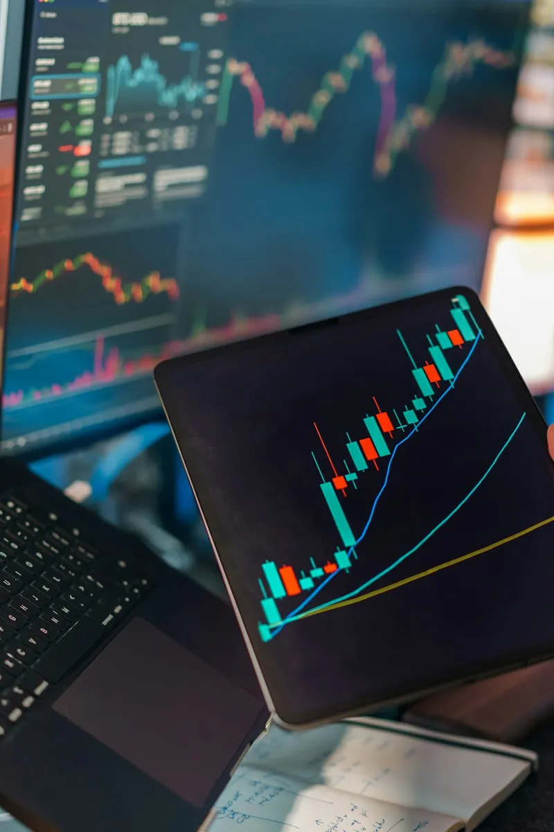 Trader holding a tablet showing a candlestick chart with an uptrending moving average line, laptop and monitor with market data blurred in the background
