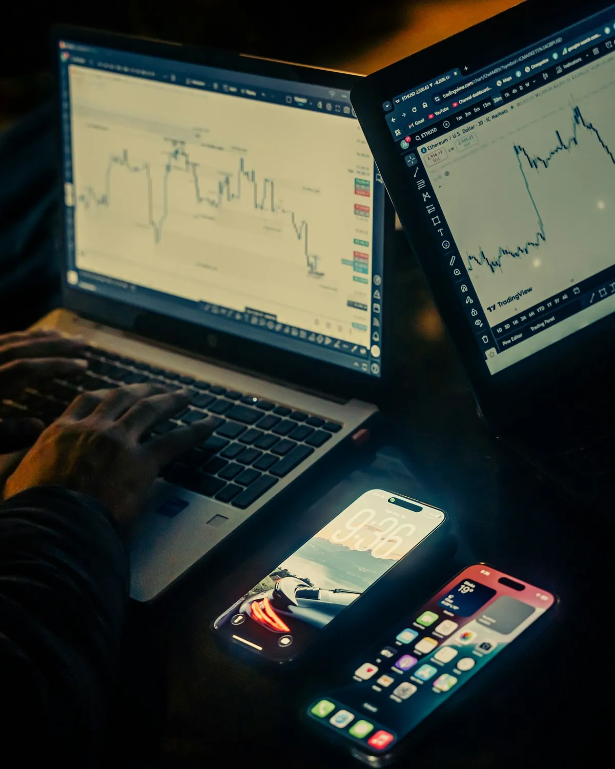 Trader's desk at night with a laptop showing a candlestick price chart and a tablet displaying a TradingView chart, phones resting nearby