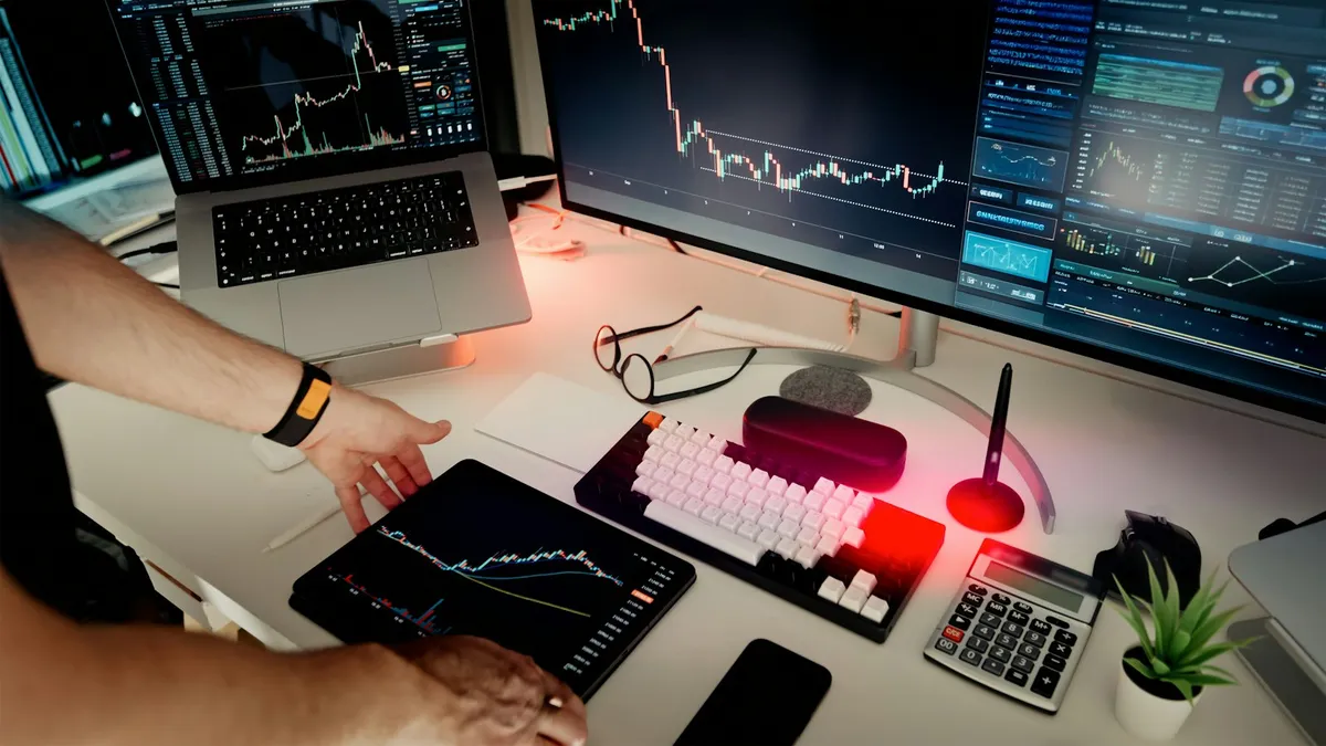Overhead view of a trader's desk with a laptop, monitor, and tablet all displaying candlestick trading charts, beside a keyboard, glasses, and calculator