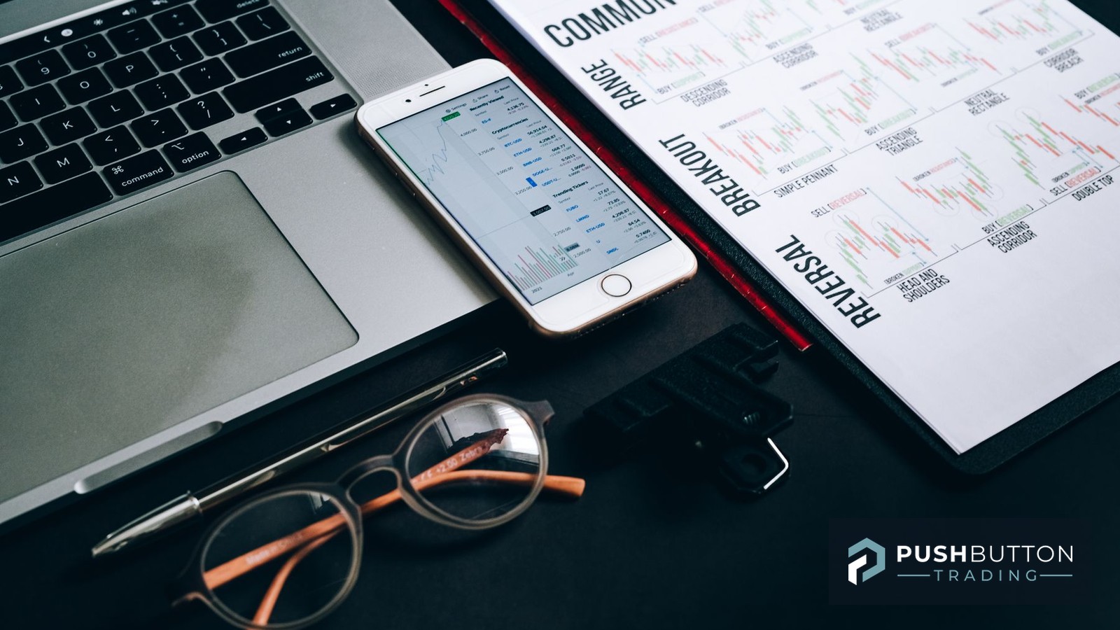 A laptop, a phone showing live tickers and a printed reference sheet of range, breakout and reversal chart patterns on a trader's desk